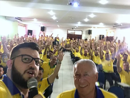 Mauro Fagotti com microfone em frente a um auditório cheio de funcionários de uniforme, todos de braços levantados